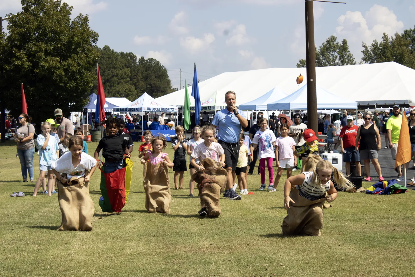Children participating in a sack race on a grassy field during a sunny outdoor community event with tents and spectators.