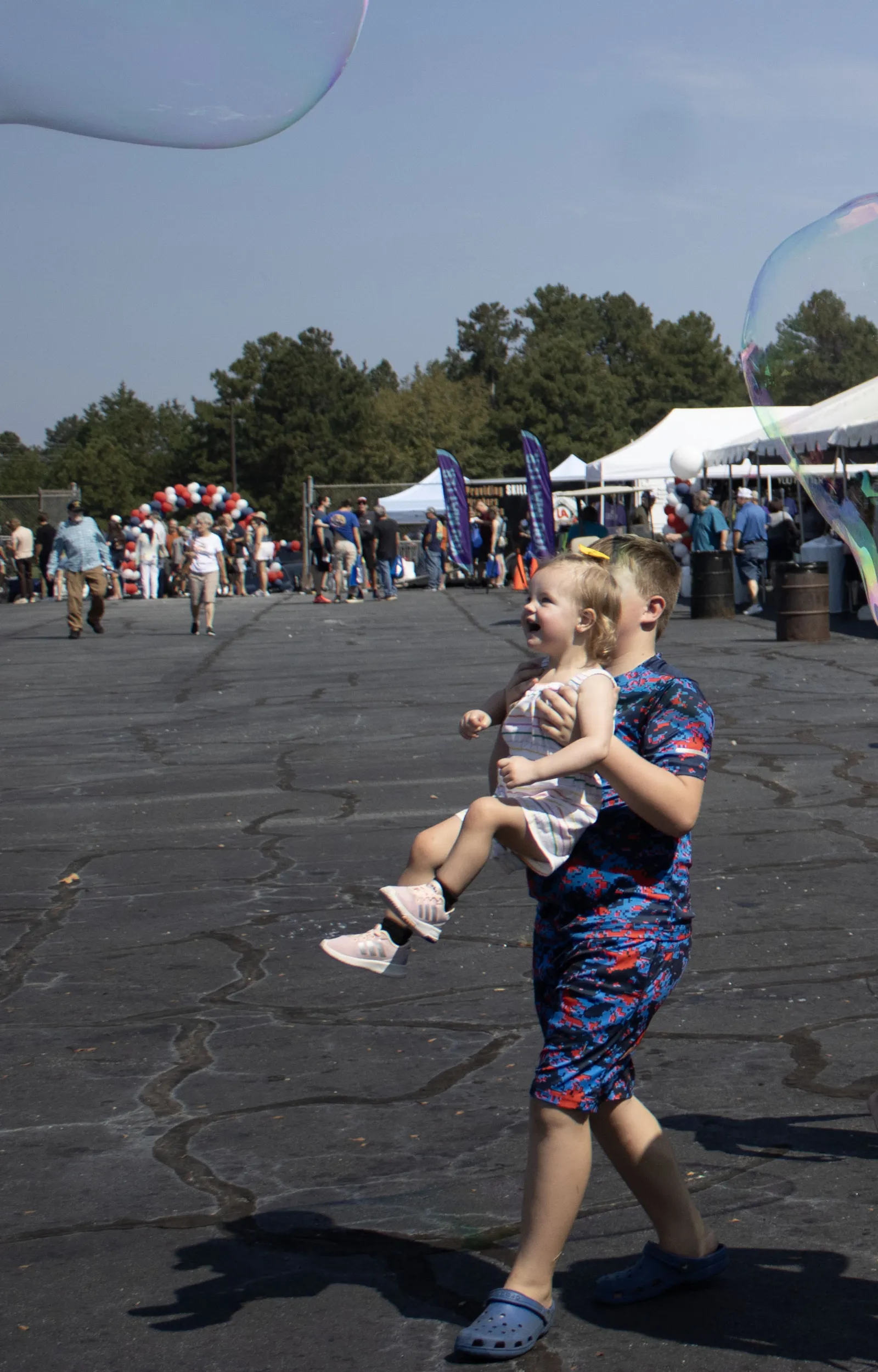 Boy carrying a smiling toddler girl outdoors with tents and people in the background on a sunny day.