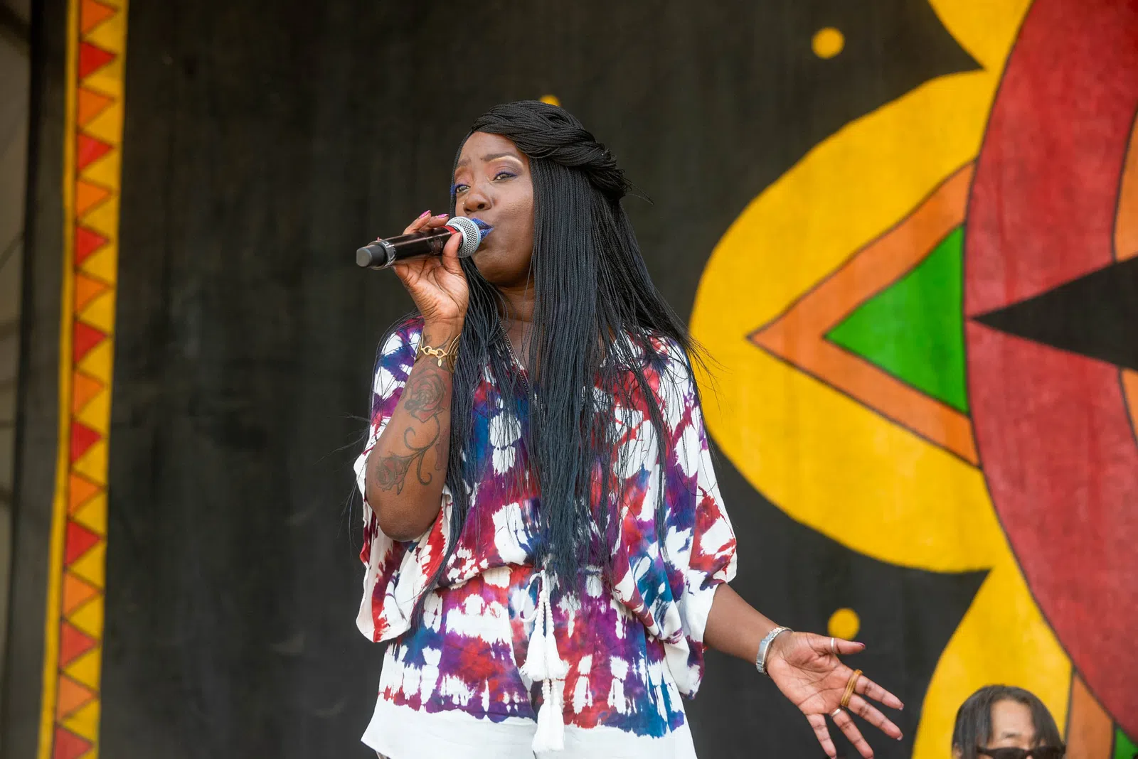 Female singer with long black hair and tattoo sings into microphone on stage with colorful backdrop.