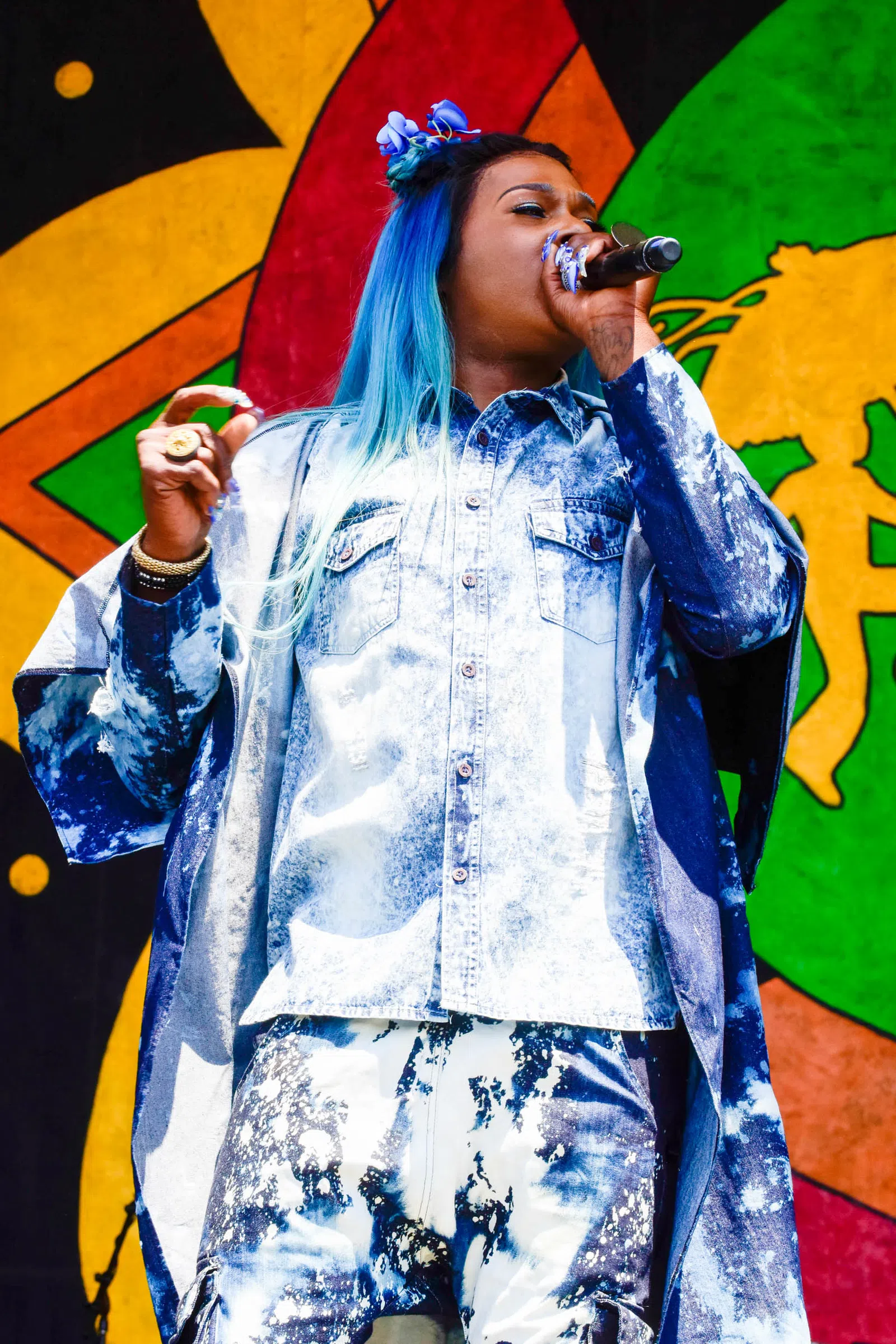 Singer with blue ombre hair and acid-wash denim outfit performing on colorful stage with microphone.