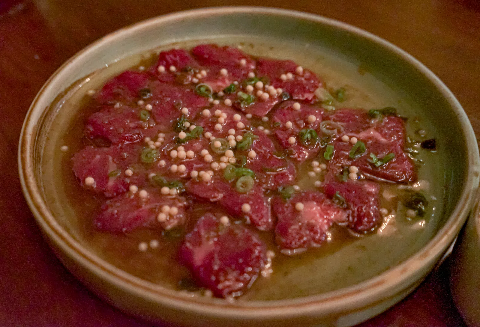 Plate of thinly sliced raw beef carpaccio topped with mustard seeds and chopped scallions in sauce.