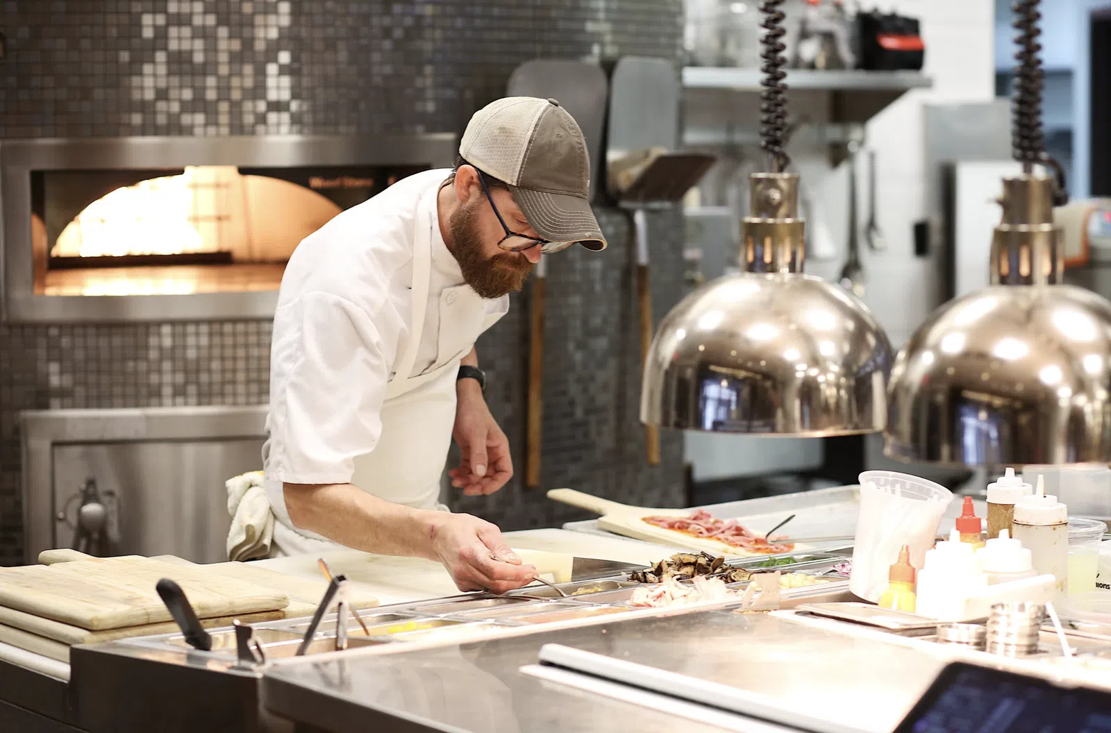 Chef in white apron adding toppings to pizzas in a professional kitchen with a wood-fired oven in the background.