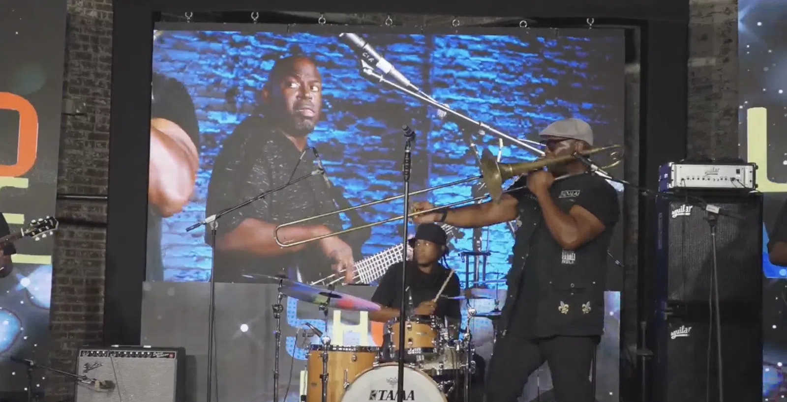 Jazz band performing live with trombone player in foreground and drummer and bassist on screen behind.