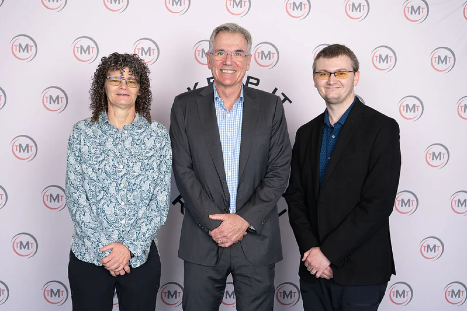 Three people posing professionally in front of a backdrop with TMT logos, dressed in business and casual attire.