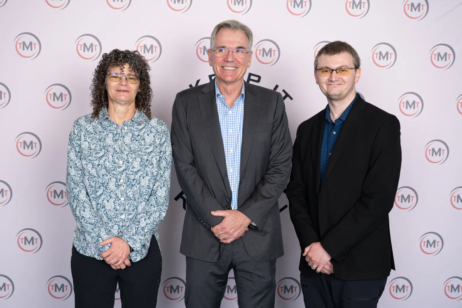 Three professionally dressed people standing smiling in front of a TMT branded backdrop at an event.