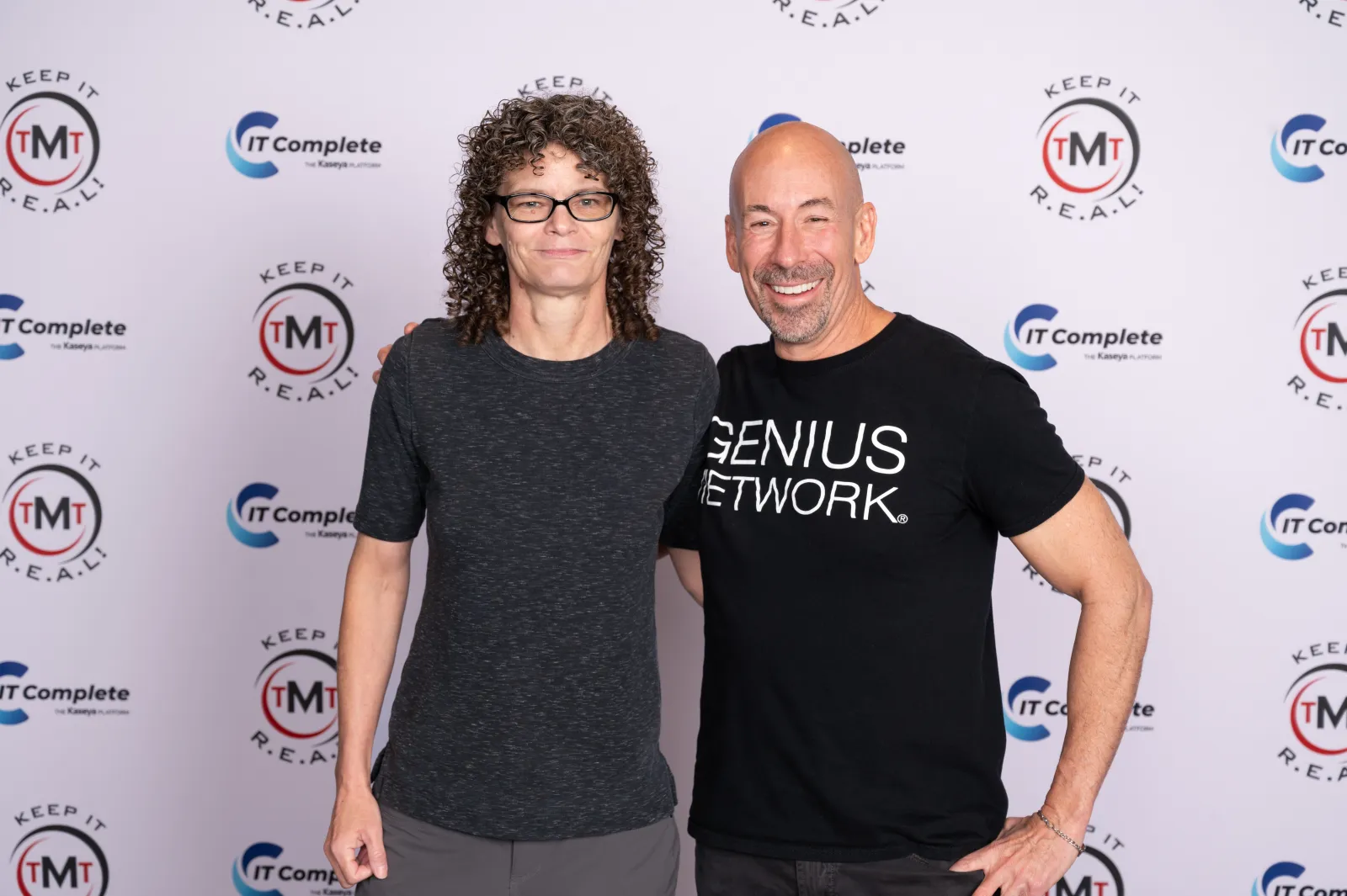 Two smiling adults posing in front of a branded backdrop with logos and slogans for IT Complete and TMT Keep It Real