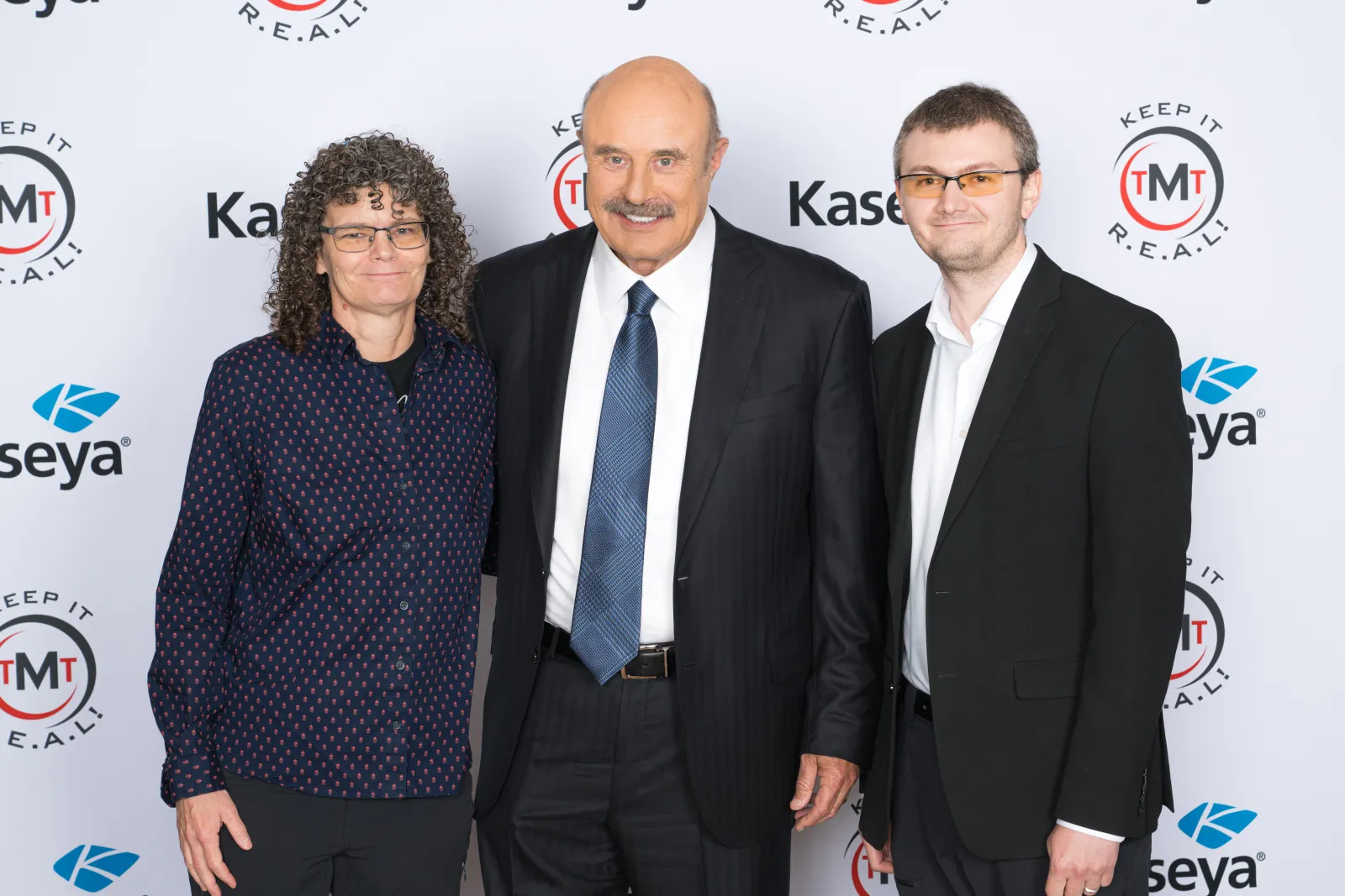 Three professionally dressed individuals posing at a media event with branded backdrop logos.