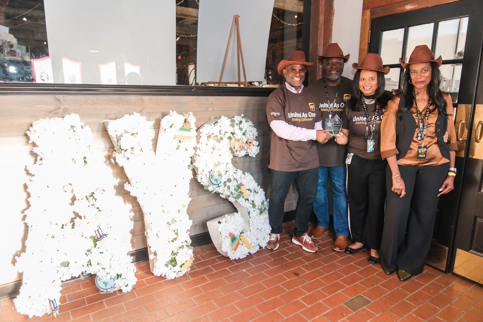 Four adults in brown shirts and cowboy hats posing next to large floral letters spelling AYS indoors.