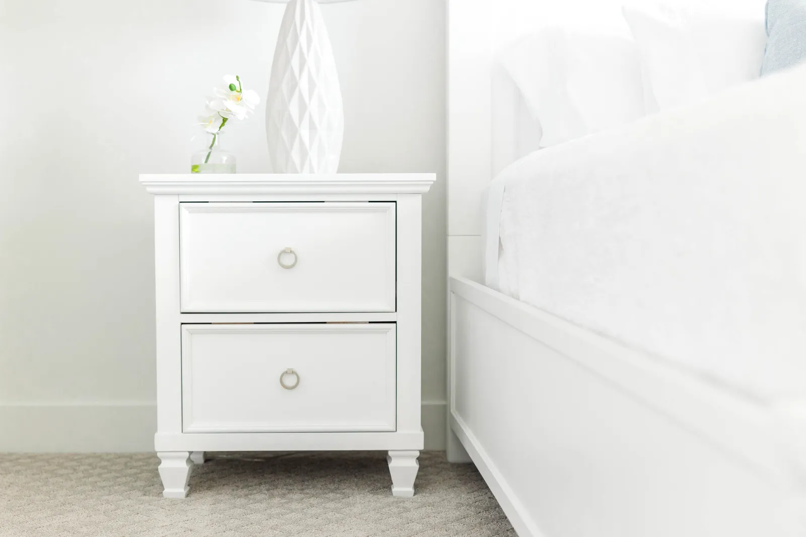 white end table with white flowers in a glass vase on top, next to a white bed set on a white bed frame on a white textured carpet edges