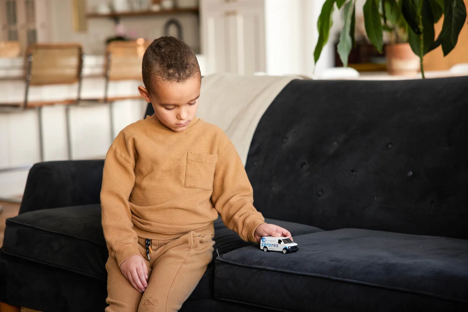 a little boy in a tan sweat suit sitting on a suede couch playing with a small Zerorez toy van