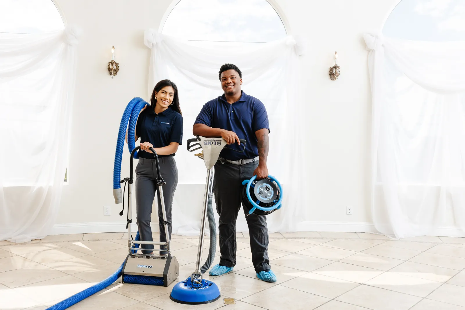 two Zerorez® technicians, female on the left and male on the right, holding various professional tie and grout cleaning equipment and tools and hoses and fans