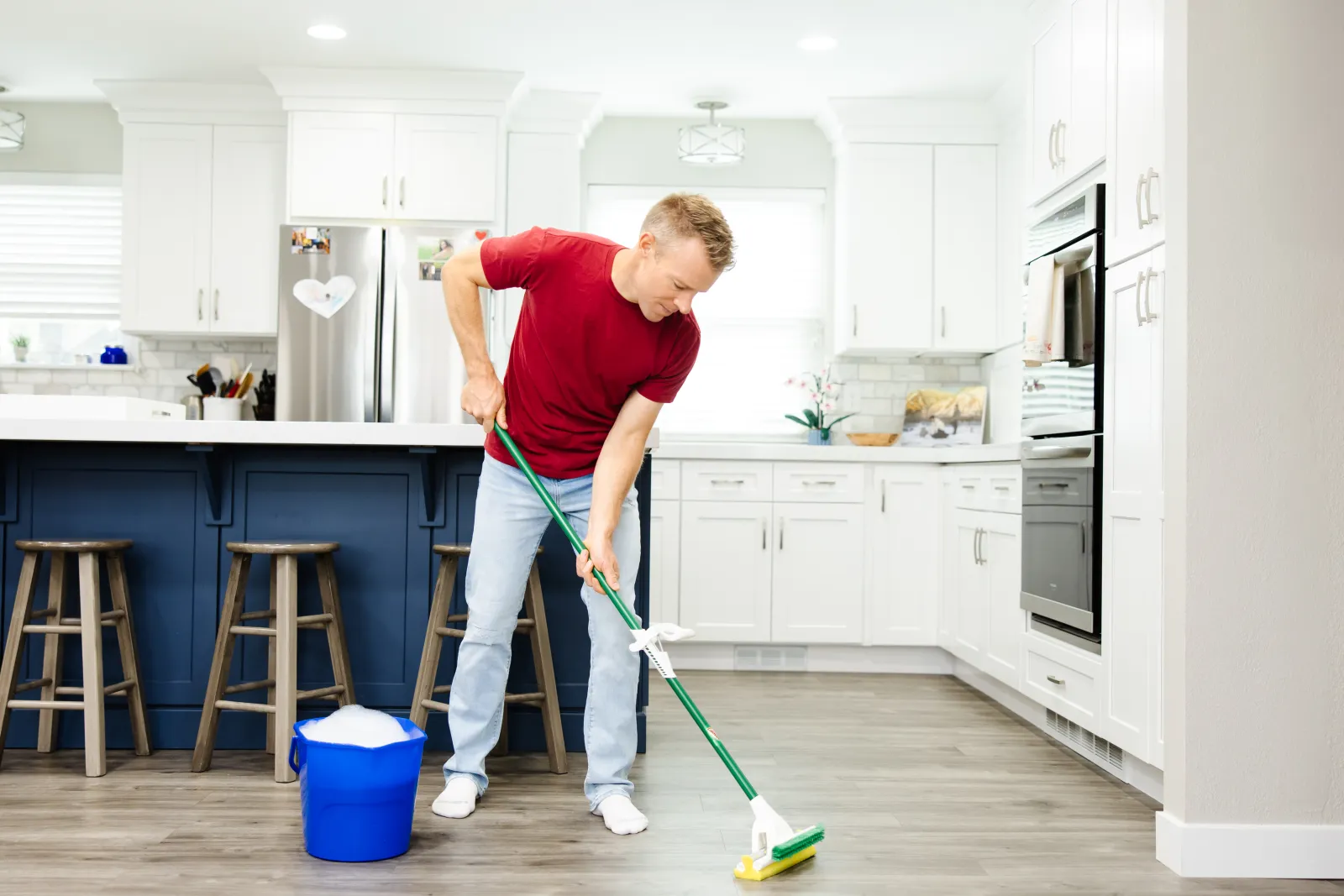 Man in a red shirt and blue jeans mopping a kitchen hardwood floor with soapy water in a blue bucket