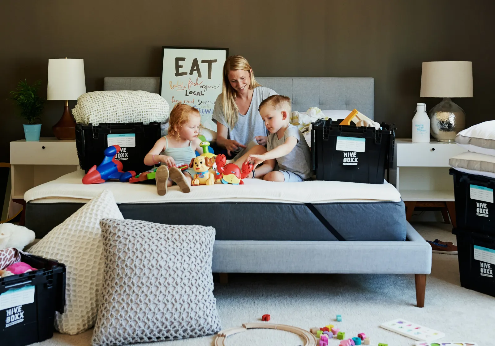 mom with two kids playing on an unmade bed with toys and boxes on the bed, as they are surrounded by moving boxes and disarray as they try to move into their new home with carpet
