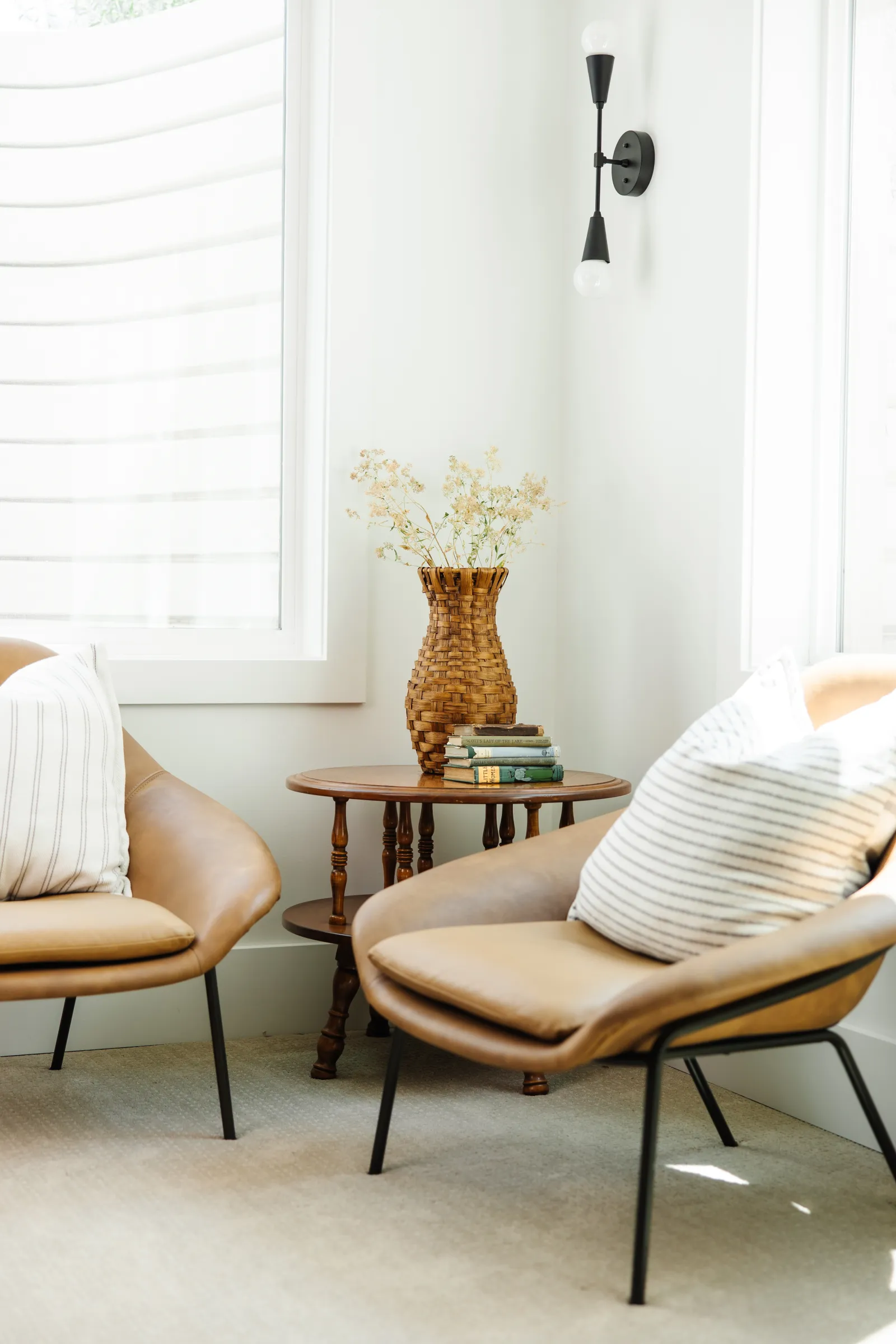 two nice leather chairs and a side table with books and a wicker vase on it in the corner of a white room with bright windows