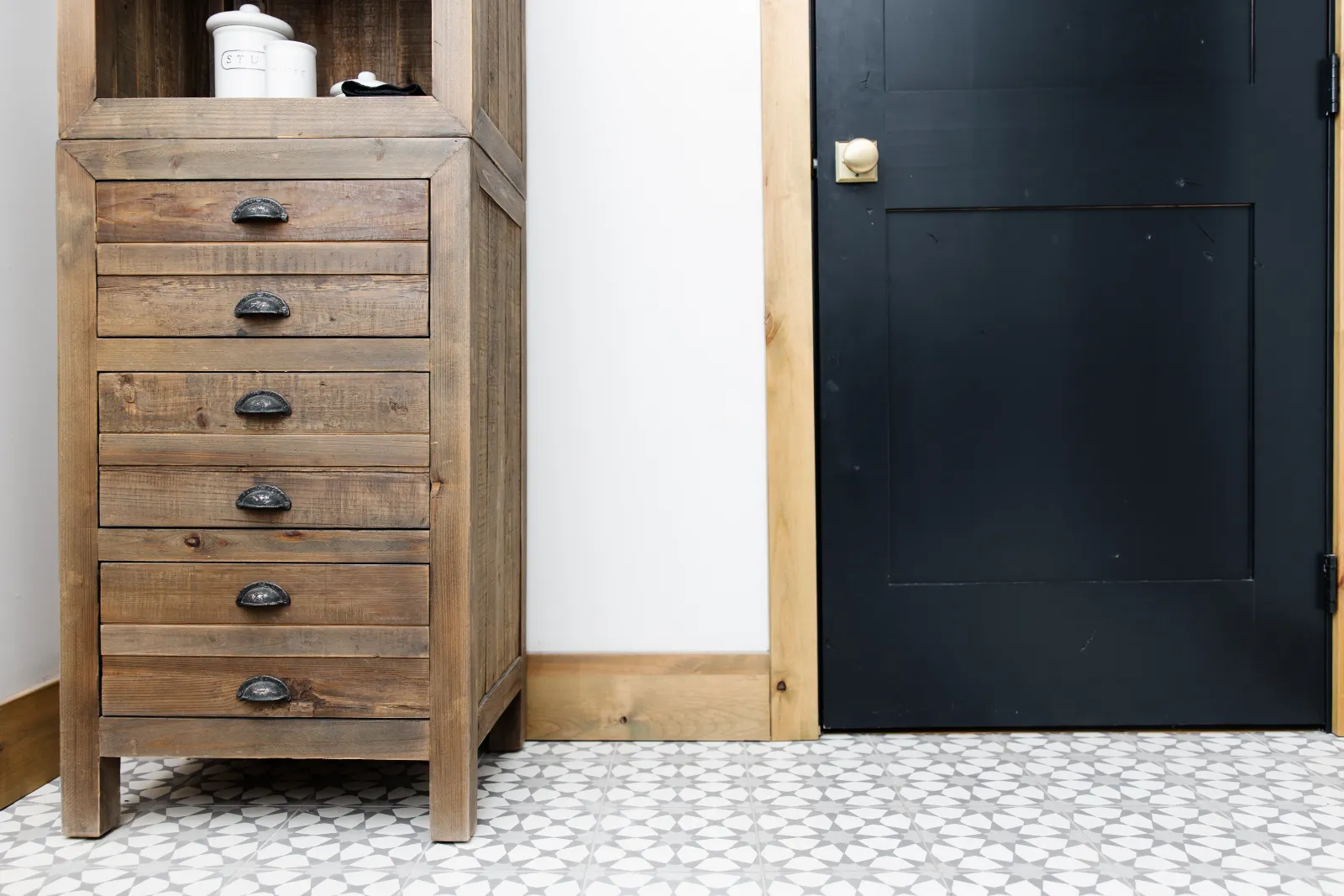 narrow tall wood dresser on a white patterned cold tile floor and a black door