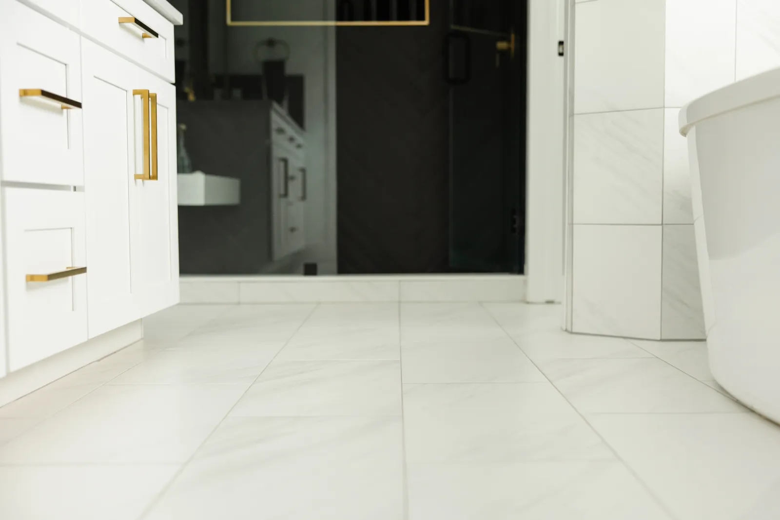 white bathroom tiles on floor and walls with white cabinets with gold handles and a dark black tiled shower behind glass