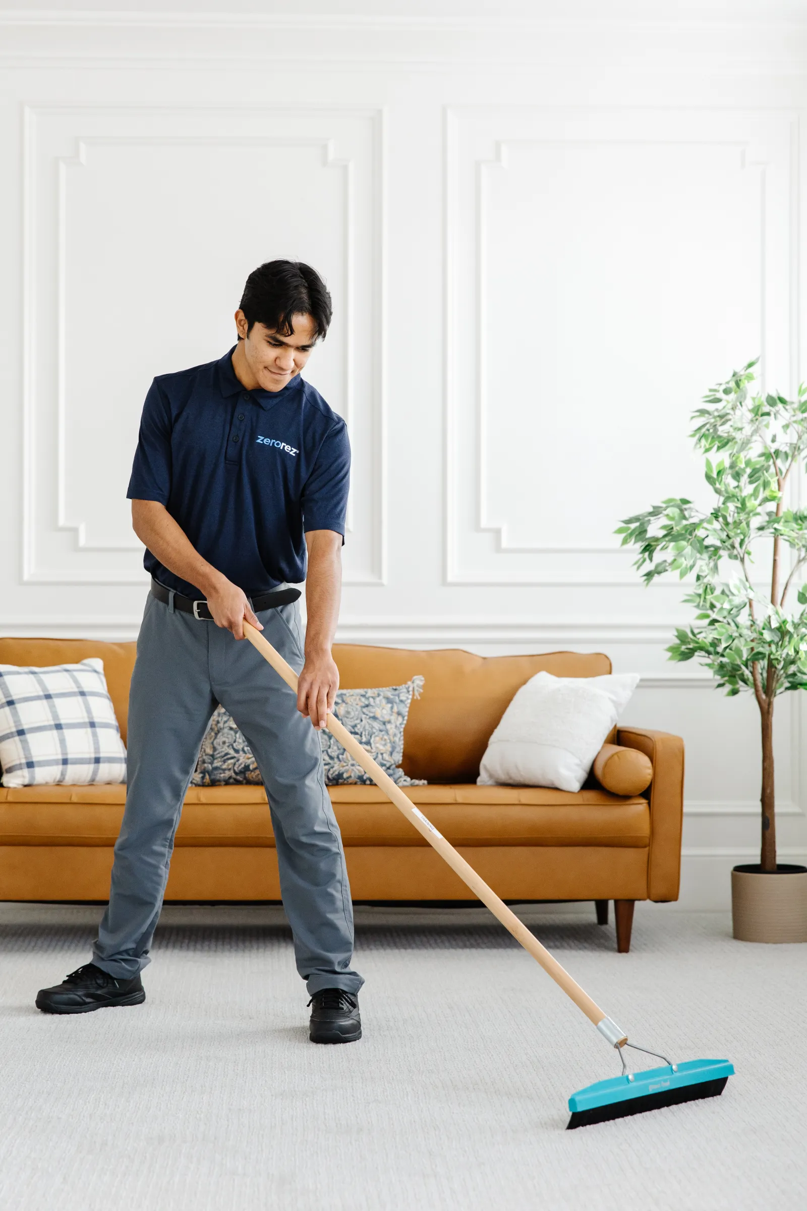 Male Latino Zerorez carpet cleaning technician standing in a living room using a carpet rake to work in the spray carpet fiber protector evenly into the light gray carpet