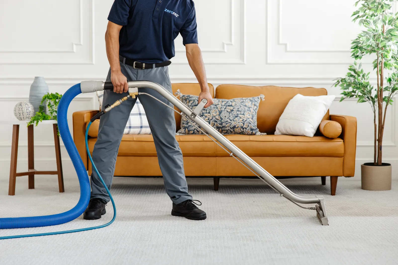 professional male Zerorez technician cleaning a light gray carpet in front of a tan leather couch to help remove dents in carpet from furniture