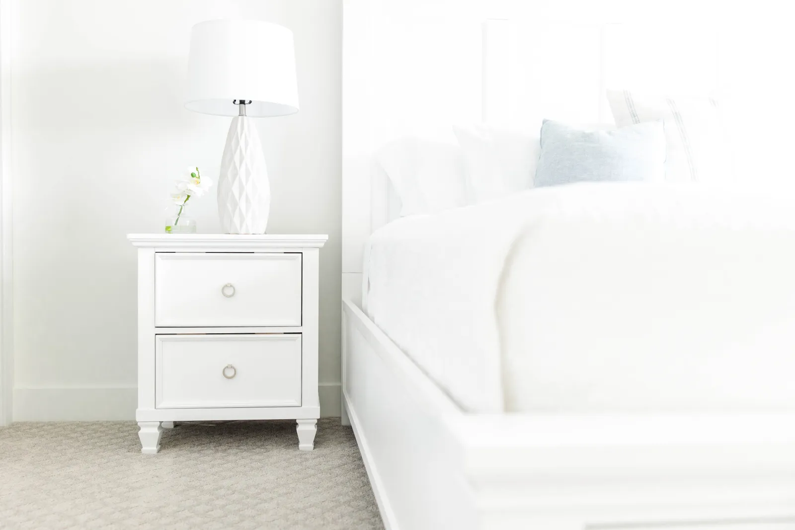 white end table with white lamp on it next to a white bed set and bedding on a light gray carpet