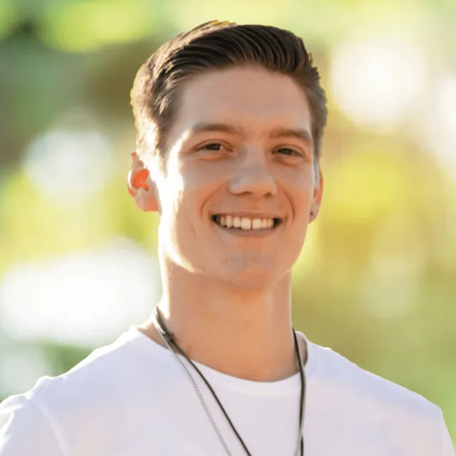 Smiling young man with short hair wearing a white shirt and necklace outdoors in soft sunlight.