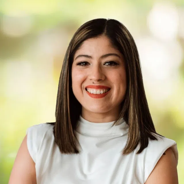 Smiling woman with straight brown hair wearing a white sleeveless top against a blurred green background