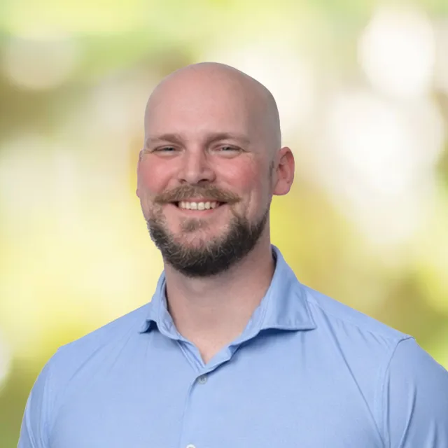Smiling bald man with a beard wearing a light blue button-up shirt with a soft nature background