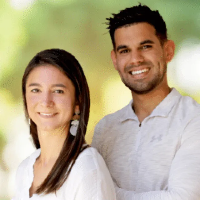 Smiling young man and woman standing outdoors with blurred green background, both wearing light-colored tops.