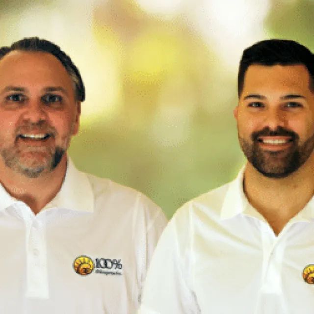 Two men smiling wearing white polo shirts with 100% Chiropractic logo on blurred background