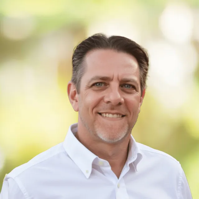 Smiling middle-aged man with short brown hair and a trimmed beard wearing a white shirt outdoors.