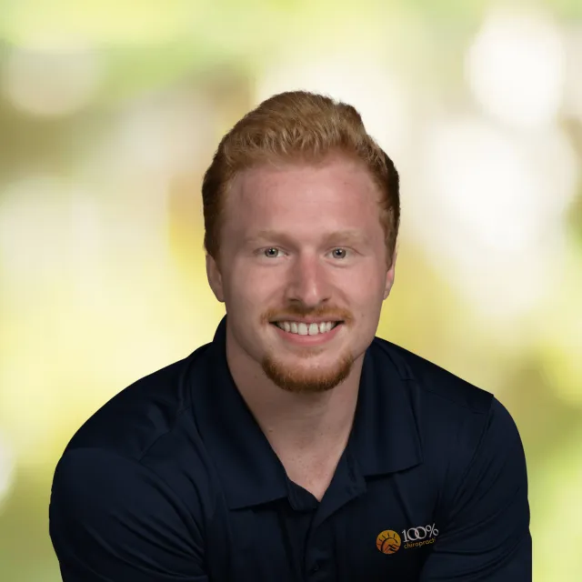 Smiling young man with red hair wearing navy blue polo shirt with 100% Chiropractic logo on a blurred green background