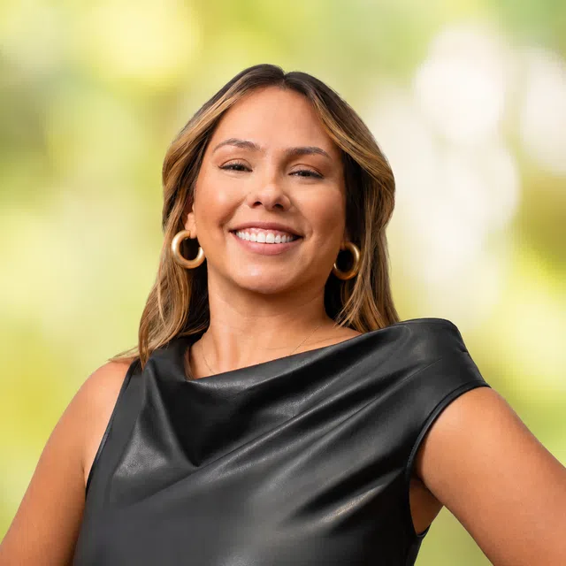 Smiling woman with wavy hair wearing hoop earrings and a black sleeveless top against a blurred green background