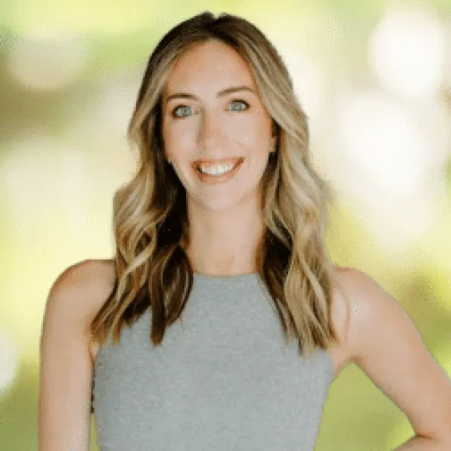Smiling woman with blonde wavy hair wearing a sleeveless gray top against a blurred light background