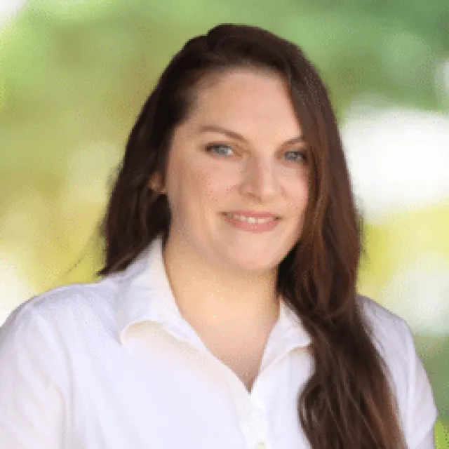 Smiling woman with long brown hair wearing a white shirt standing outdoors with blurred green background