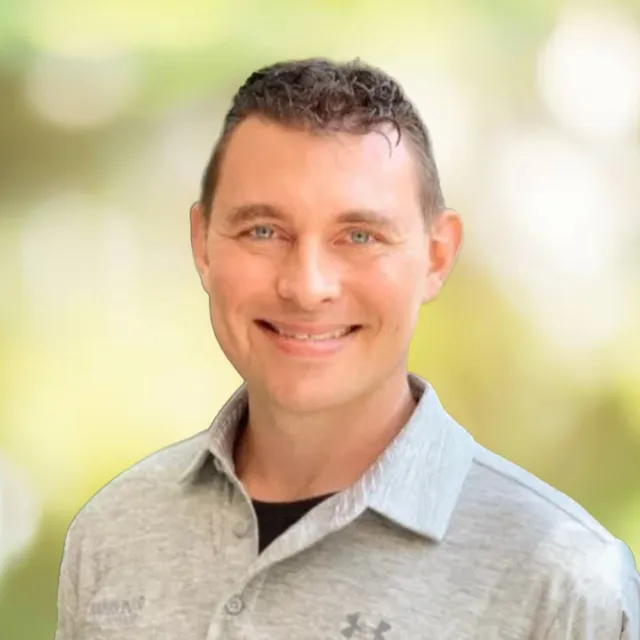 Smiling man with blue eyes wearing a light gray collared shirt in a softly blurred outdoor setting.