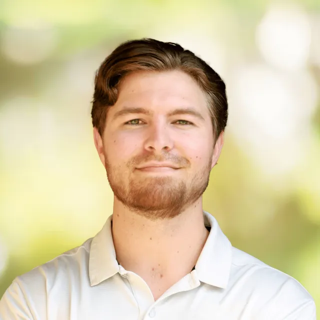 Confident young man with short brown hair and beard wearing a white polo shirt against a blurred outdoor background