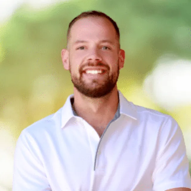 Smiling man with beard wearing a white collared shirt standing outdoors with blurred greenery background