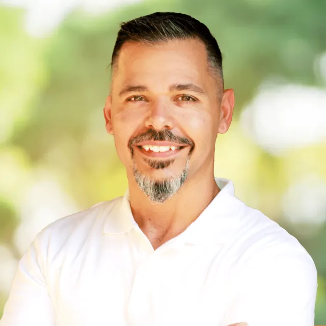 Smiling man with trimmed beard wearing a white polo shirt standing outdoors with blurred greenery background.
