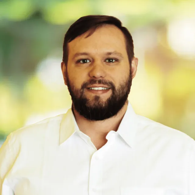 Smiling man with beard wearing a white collared shirt against a blurred green and yellow background