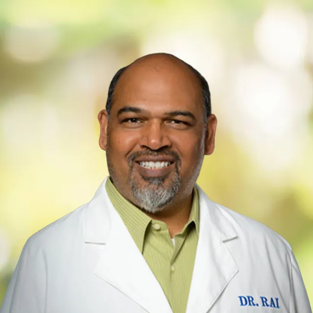 Smiling male doctor with beard wearing a white lab coat and green shirt on blurred bright background