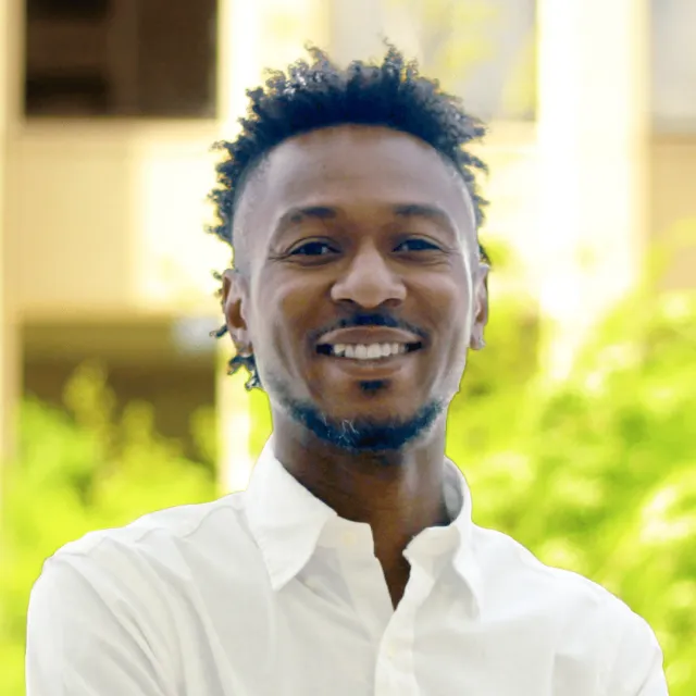 Smiling man with short curly hair wearing a white shirt standing outdoors with blurred greenery background