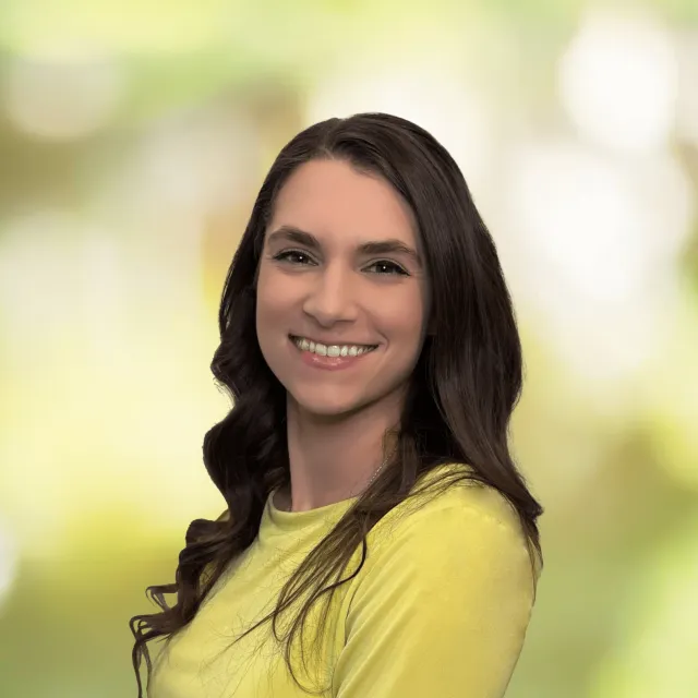 Young woman with long dark hair smiling in a yellow top against a soft green blurred background