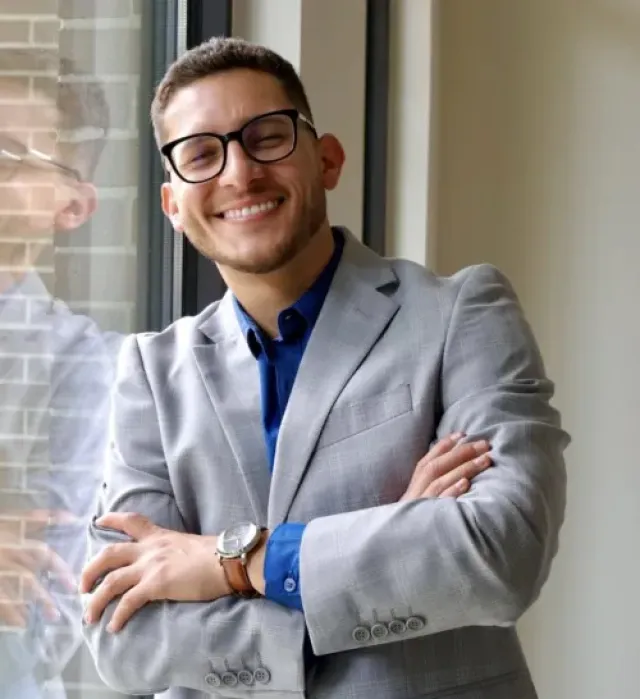 Smiling young man in glasses wearing a gray blazer and blue shirt standing with arms crossed by a window.
