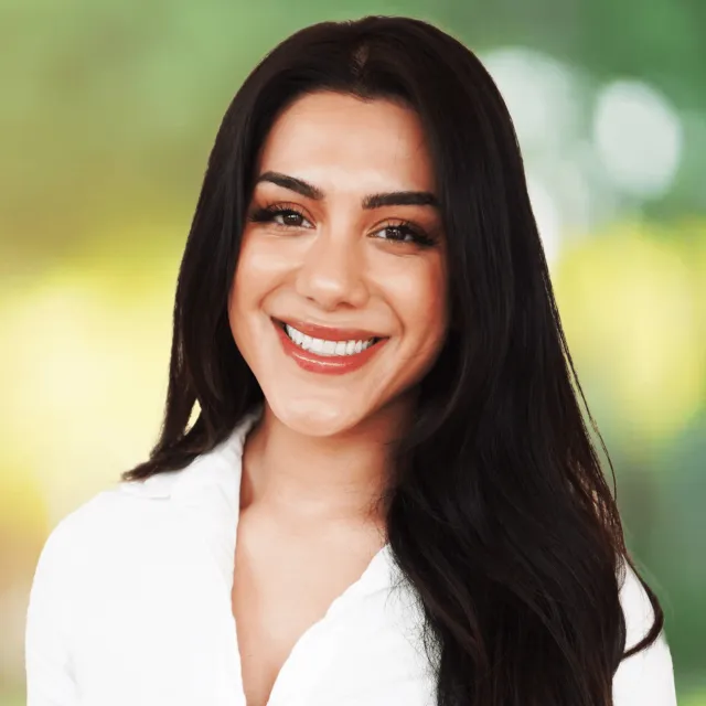 Smiling young woman with long dark hair wearing a white shirt against a blurred outdoor background.
