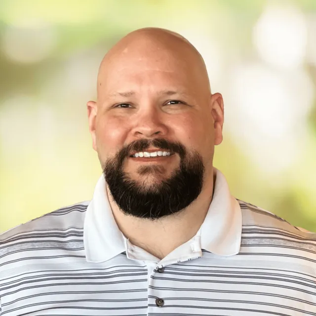 Smiling bald man with a dark beard wearing a white and gray striped polo shirt, blurred green background.