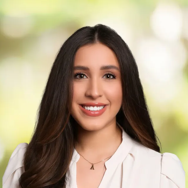 Smiling woman with long dark hair wearing a white blouse and letter A necklace against blurred green background
