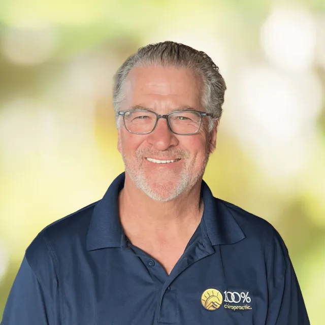 Smiling middle-aged man with gray hair and glasses wearing a navy blue 100% Chiropractic polo shirt