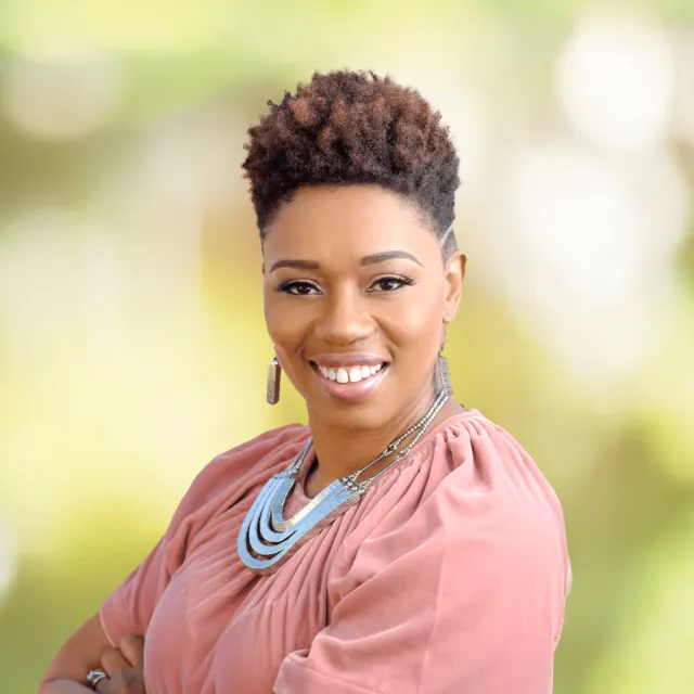Smiling woman with short curly hair wearing a pink blouse and silver necklace against a blurred green background