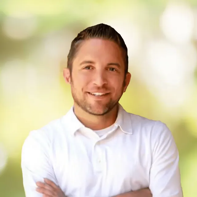 Smiling man in white button-up shirt standing with arms crossed against a blurred green and white background