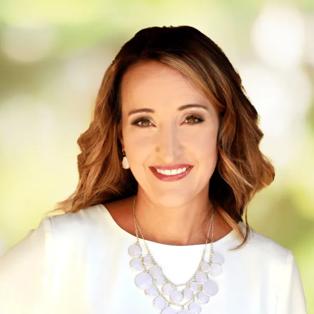 Smiling woman with wavy brown hair wearing a white top and statement necklace against a blurred green background