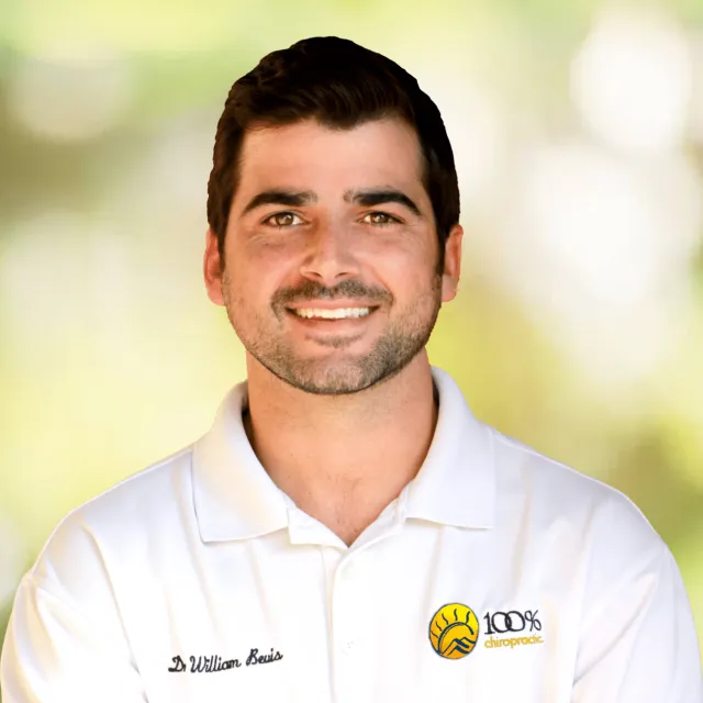 Smiling male chiropractor wearing a white polo shirt with 100% Chiropractic logo on blurred green background